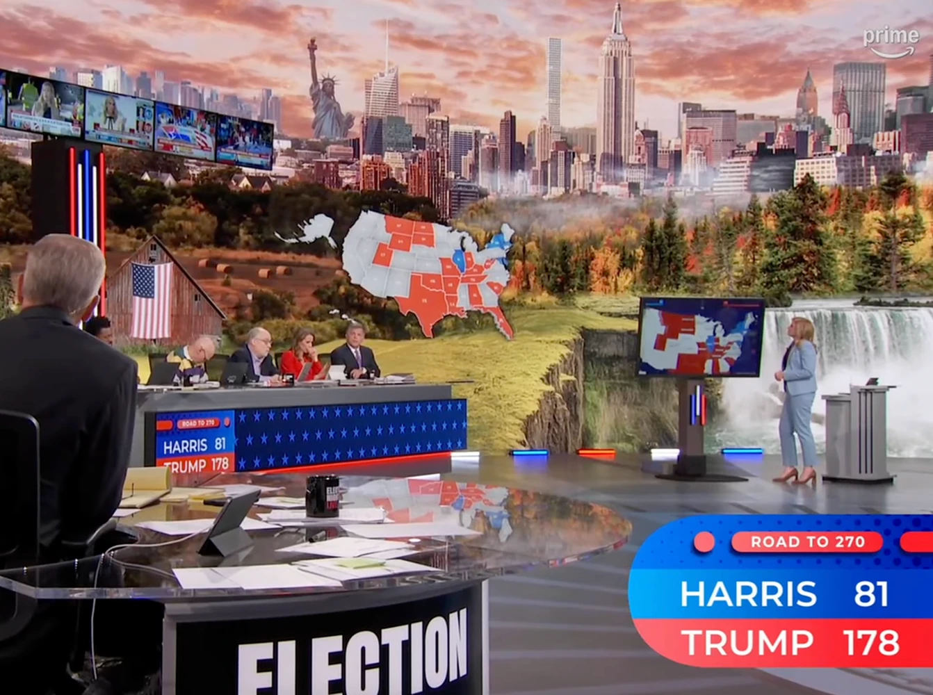 A large television news studio shows anchors seated at a desk while a presenter stands near a screen displaying a color-coded map of the United States with electoral vote counts for presidential candidates.